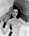 Ann Miller frolics in the studio for Ned Scott's camera to support her role as Edithea Alden, college girl moonlighting as a nightclub dancer in Director Arthur Dreifuss’ musical comedy, "Eadie Was A Lady", 1945. Large hats often figure in Ned Scott's portraits of Hollywood personalities and commercial modeling subjects. The caption on the back of this print sports the humorous quip, "Who's looking at a hat when Ann Miller is under it"