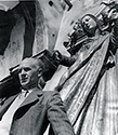 With arms outstretched , a bas-relief statue of the Virgin Mary gathers protectively behind Ernie Pyle as he poses on the set of The Story of G.I. Joe. Ned Scott was making a hopeful statement with this prosaic pose. Keep Ernie Safe. In October and November of 1944 when this photograph was taken, no one could have predicted the tragic fate which would befall Ernie has he ventured to the Pacific Theater in 1945 on non-combatant duty as a U.S. War Correspondent. 