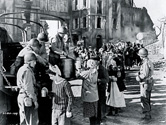 In this scene for the film Story of G.I. Joe, army cooks flanked by armed soldiers serve hot food to refugees whose homes were destroyed in a recent battle. One of the major objectives of Director William Wellman was to convey battlefield authenticity, complete with scenes such as these which occurred in real life in Southern Italy and Northern Africa on a regular basis as the Allies advanced in their quest to oust the Germans and Italians. 