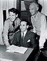 Lester Cowan, seated, is flanked on the left by Captain Burgess Meredith, recently furloughed from active duty by the War Department for his role in The Story of G.I. Joe, and Ernie Pyle, U.S. War Correspondent from Scripps-Howard News Service. In the film, Meredith played the the role of Ernie Pyle. Ned Scott took this photograph in an office at Selznick International Studios in Culver City, California.